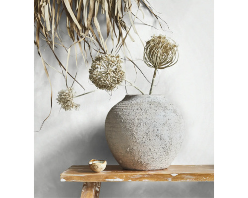 Ronde keramieken vaas met ruwe textuur en droogbloemen op een houten bankje voor een lichte wand.