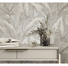 Licht vliesbehang met groot tropisch bladpatroon en bloemen achter een dressoir met boeken en een bloemenvaas.