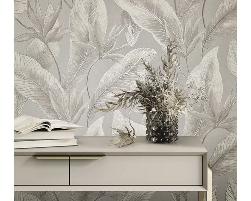 Licht vliesbehang met groot tropisch bladpatroon en bloemen achter een dressoir met boeken en een bloemenvaas.