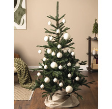 Groene kerstboom versierd met witte kerstballen in verschillende maten en een stoffen boomrok in een moderne woonkamer.