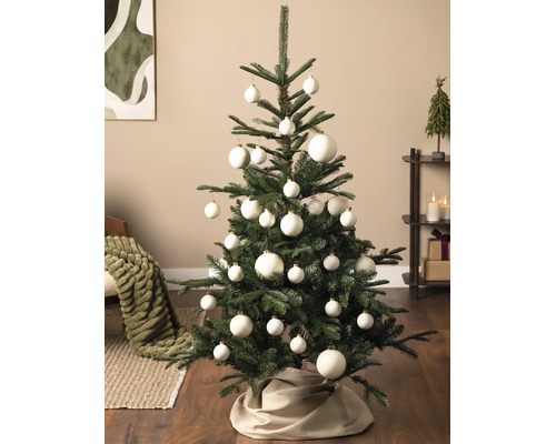 Groene kerstboom versierd met witte kerstballen in verschillende maten en een stoffen boomrok in een moderne woonkamer.