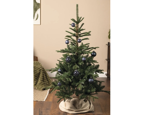 Groene kerstboom versierd met blauwe kerstballen met patronen op een houten vloer met een beige kerstboomrok.