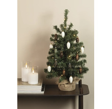 Kleine kunstkerstboom met dennenappelhangers, lichtsnoer en jutiezak naast twee witte kaarsen in glas op een stapel tijdschriften.