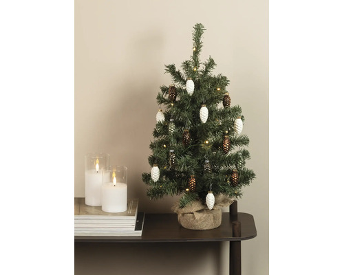 Kleine kunstkerstboom met dennenappelhangers, lichtsnoer en jutiezak naast twee witte kaarsen in glas op een stapel tijdschriften.