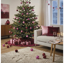Feestelijk versierde kerstboom in een lichte woonkamer met roze en bordeauxrode ballen, lichtsnoer, cadeaus en een bank.