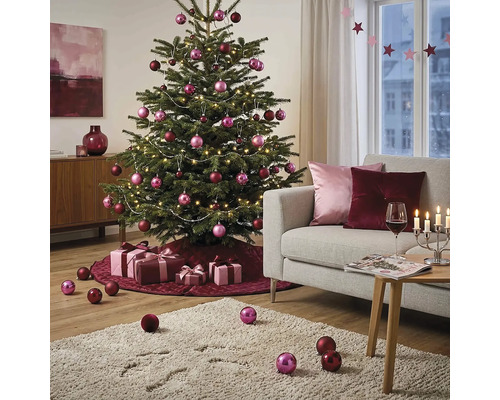 Feestelijk versierde kerstboom in een lichte woonkamer met roze en bordeauxrode ballen, lichtsnoer, cadeaus en een bank.