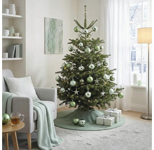 Versierde kerstboom met kerstballen en lichtjes in een lichte woonkamer naast een fauteuil met gebreide deken en geschenken.