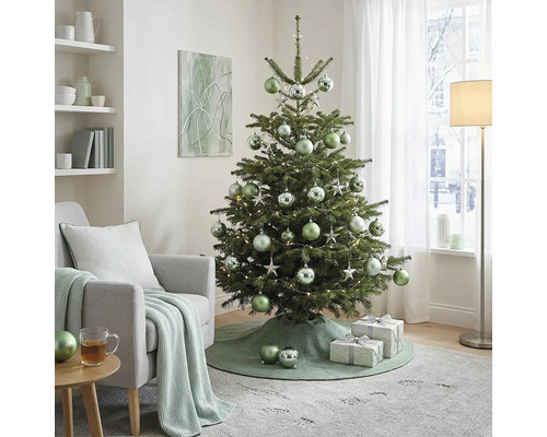 Versierde kerstboom met kerstballen en lichtjes in een lichte woonkamer naast een fauteuil met gebreide deken en geschenken.