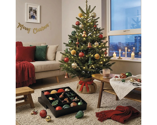 Gedecoreerde kerstboom met verlichting in een woonkamer met bank en een doos vol kerstversiering op het tapijt.