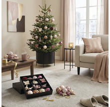 Feestelijk versierde kerstboom in een metalen standaard in een lichte woonkamer met bank, salontafel en kerstballen in een doos.