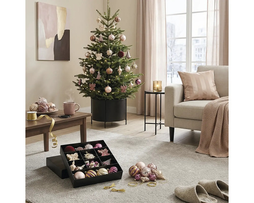 Feestelijk versierde kerstboom in een metalen standaard in een lichte woonkamer met bank, salontafel en kerstballen in een doos.