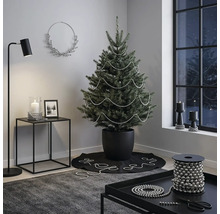 Modern ingerichte kamer met een kerstboom in een zwarte pot, gedecoreerd met zilveren parelslingers, een bijzettafel en een vloerlamp.