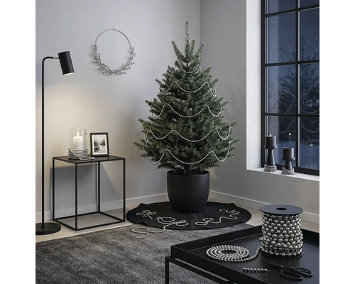 Modern ingerichte kamer met een kerstboom in een zwarte pot, gedecoreerd met zilveren parelslingers, een bijzettafel en een vloerlamp.