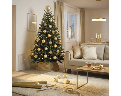 Feestelijk versierde kunstkerstboom met gouden kerstballen en lichtjes in een lichte woonkamer met bank en salontafel.
