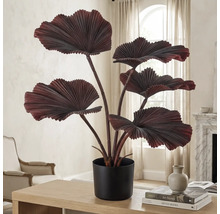 Kunstplant met grote, geplooide waaierbladeren in een zwarte pot op een houten tafel in een lichte, moderne woonkamer.