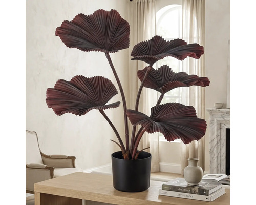 Kunstplant met grote, geplooide waaierbladeren in een zwarte pot op een houten tafel in een lichte, moderne woonkamer.