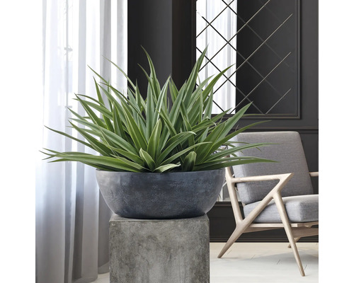 Kunstplant met lange, puntige bladeren in een donkergrijze plantenschaal op een betonnen sokkel in een modern ingerichte kamer.