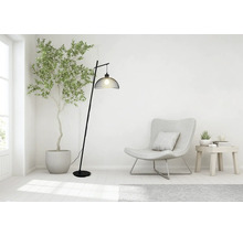Moderne booglamp van zwart metaal met een kap van gaas in een lichte woonkamer met fauteuil en kamerplant.