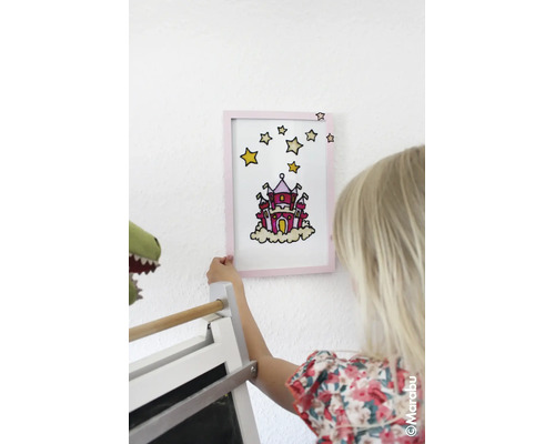Een kind houdt een met glasverf gemaakte afbeelding van een kasteel in een lijst tegen de muur. Marabu Logo.