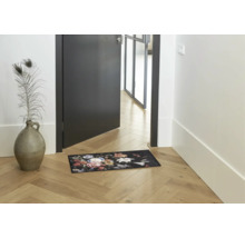 Deurmat met bloemenpatroon in een lichte hal met houten vloer naast een deur en een vaas.