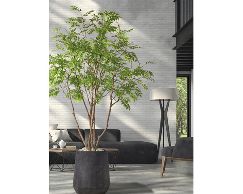 Grote kunstboom in een donkere plantenbak in een moderne woonkamer met een witte bakstenen muur, een donkere bank en een vloerlamp.