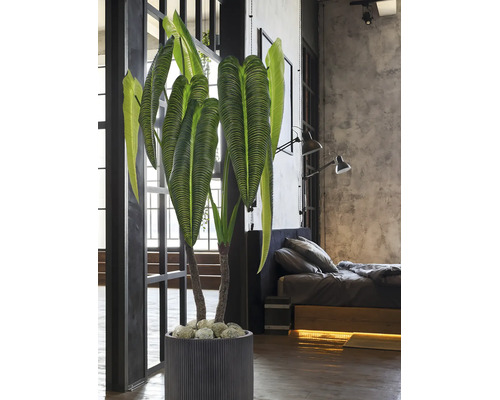 Grote kunstplant met geribbelde bladeren in een grijze pot met verticale groeven in een moderne slaapkamer in industriële stijl.