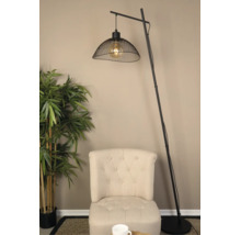 Moderne booglamp van zwart metaal met een kap van gaas, geplaatst naast een beige gecapitonneerde fauteuil en een kunstplant.