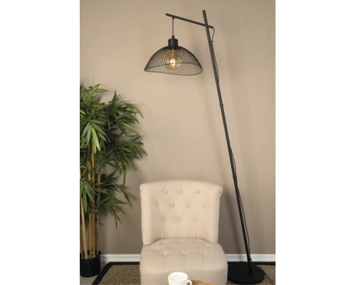 Moderne booglamp van zwart metaal met een kap van gaas, geplaatst naast een beige gecapitonneerde fauteuil en een kunstplant.