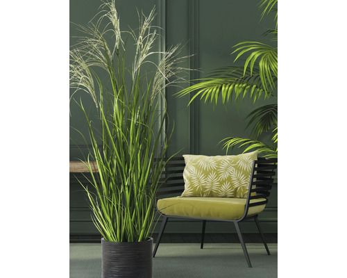 Kunstgrasplant in een zwarte geribbelde pot naast een moderne fauteuil in een groene woonruimte.