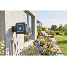 Wandslangenbox van for.q aan een buitenmuur in een verzorgde tuin met bloembedden, gazon en een ligstoel.