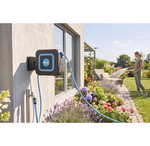Wandslangenbox met for.q Logo aan een huismuur in een lichte tuin, een vrouw besproeit bloembedden met de tuinslang.