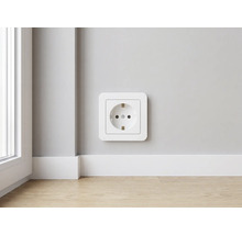 Moderne witte kunststof wandcontactdoos met randaarde aan een muur boven een witte plint en lichte houten vloer in een lichte kamer.