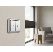 Dubbele rolluikschakelaar met pijlsymbolen omhoog en omlaag, gemonteerd op een muur in een moderne, lichte woonkamer bij een raam met jaloezieën.