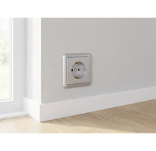 Wandcontactdoos met randaarde in zilverkleur op een lichte muur boven een witte plint in een kamer met houten vloer.