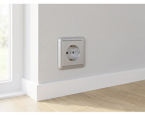 Wandcontactdoos met randaarde in zilverkleur op een lichte muur boven een witte plint in een kamer met houten vloer.