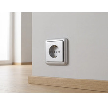 Zilverkleurige wandcontactdoos gemonteerd op een lichte muur boven een houten plint.