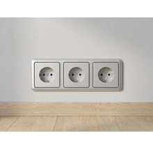 Drievoudige wandcontactdoos met randaarde en zilverkleurig frame, gemonteerd op een wand boven een houten plint.