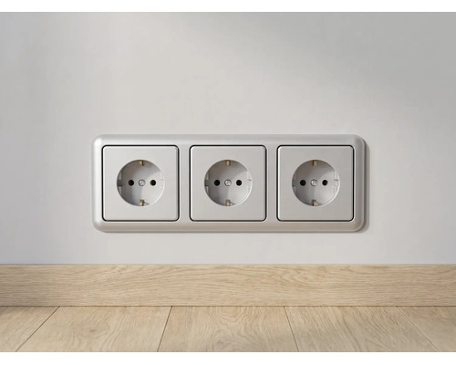 Drievoudige wandcontactdoos met randaarde en zilverkleurig frame, gemonteerd op een wand boven een houten plint.