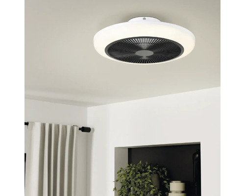 Moderne, ronde plafondventilator met LED-lichtring aan een plafond in een lichte, opgeruimde kamer met gordijnen en kamerplanten.