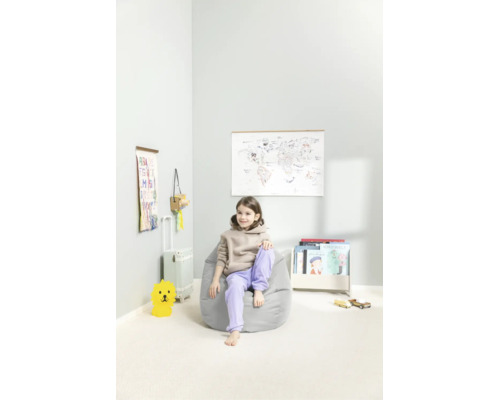 Kind zit in een lichtgrijze zitzak in een lichte, opgeruimde kinderkamer met een wereldkaart, boeken en speelgoed.