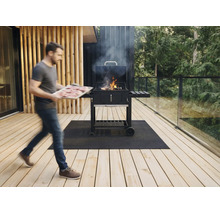 Een man draagt een plateau met vlees naar een rokende houtskoolbarbecue op een houten terras met glazen balustrade en grillmat.