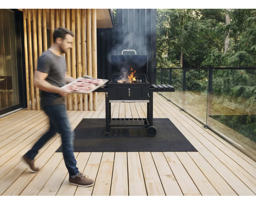 Een man draagt een plateau met vlees naar een rokende houtskoolbarbecue op een houten terras met glazen balustrade en grillmat.