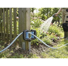 Hoekslanggeleider voor de tuin, gemonteerd aan een houten schuttingpaal, met een tuinslang en een persoon die bloemen water geeft op de achtergrond.