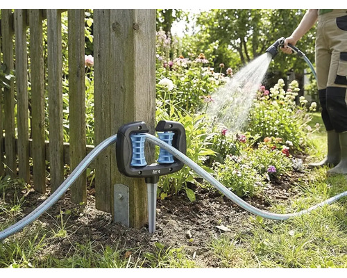 Hoekslanggeleider voor de tuin, gemonteerd aan een houten schuttingpaal, met een tuinslang en een persoon die bloemen water geeft op de achtergrond.