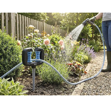 Slanggeleider in een bloembed met een tuinslang en een persoon die op de achtergrond planten water geeft.