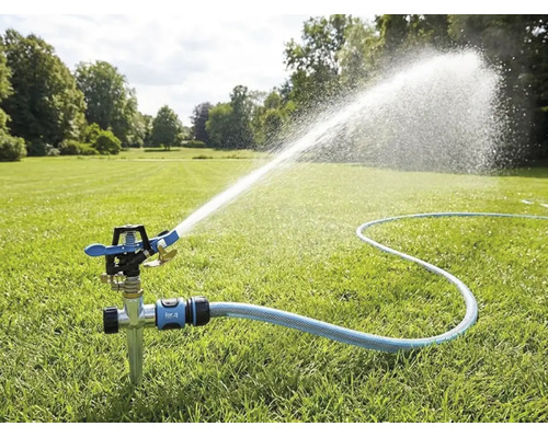 Impulssproeier met grondpen op een gazon, aangesloten op een tuinslang, sproeit een brede waterstraal voor tuinbesproeiing.