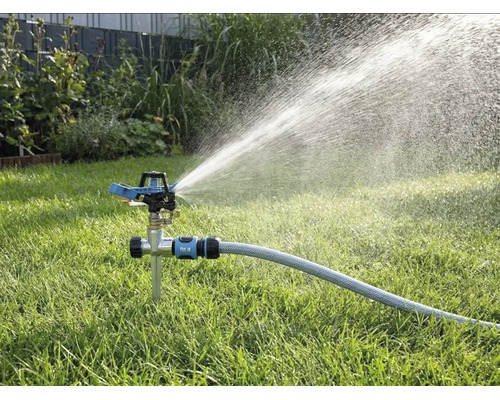 Pulsatiessproeier met grondpen op het gazon, aangesloten op een tuinslang, sproeit water in een brede straal.