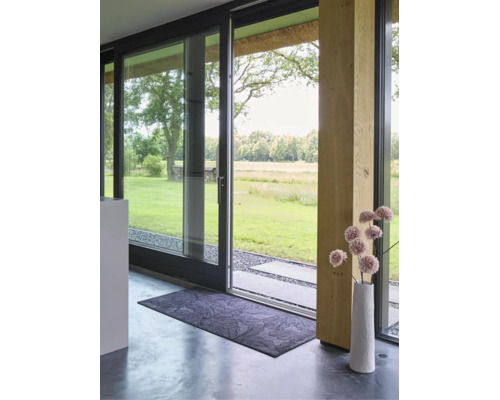 Rechthoekige deurmat met bladmotief voor een moderne glazen schuifdeur met uitzicht op de tuin en een decoratieve vaas.