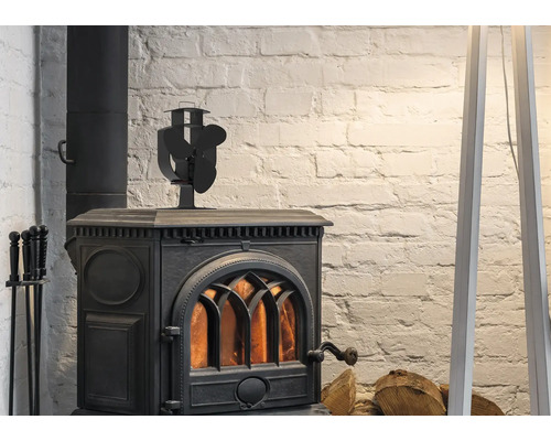 Zwarte gietijzeren houtkachel met brandend vuur, een kachelventilator bovenop en haardstel voor een witte bakstenen muur.