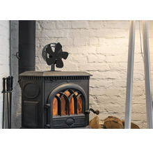 Een zwarte kachelventilator met vijf bladen op een brandende gietijzeren houtkachel voor een witte bakstenen muur met haardhout.